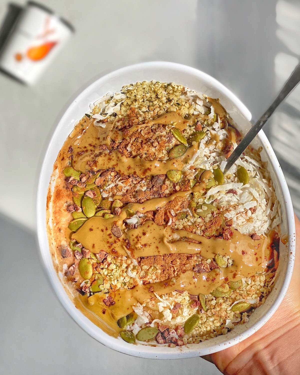 Holding white bowl with orange carrot cake smoothie bowl inside, topping with seeds and cinnamon.