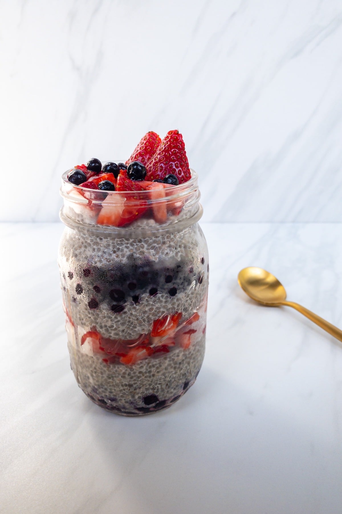 Layered chia seed pudding with strawberries and blueberries from the front with a white marble background and gold spoon