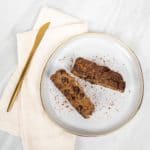 Two slices of cinnamon raisin banana bread on a white gold plate with a linen napkin and gold knife