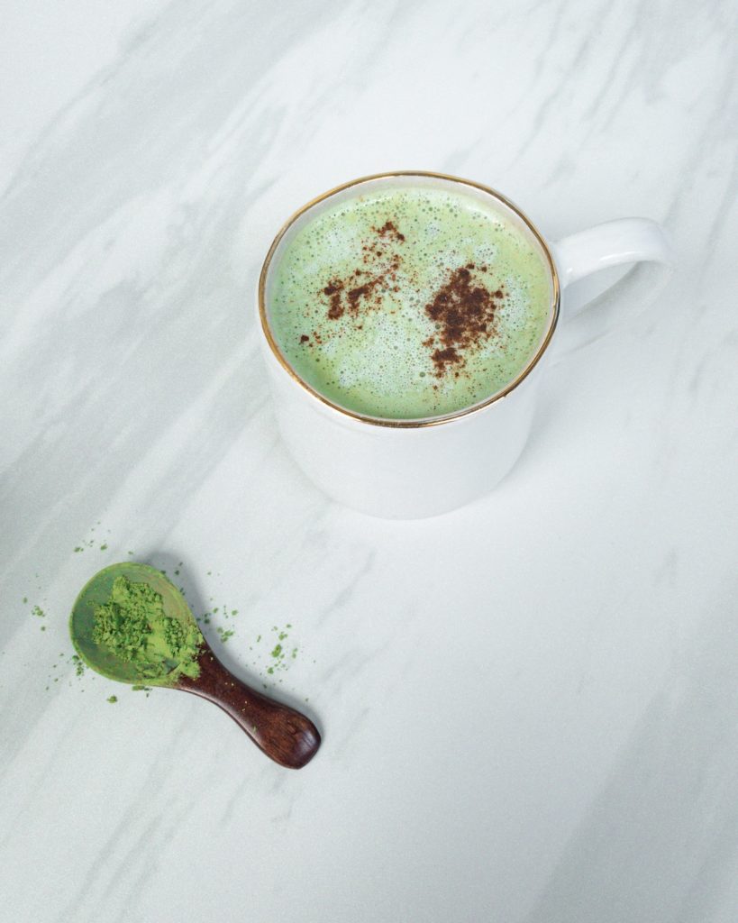 Vegan mint green matcha latte in white mug on white marble surface. Brown wooden spoon with matcha powder.