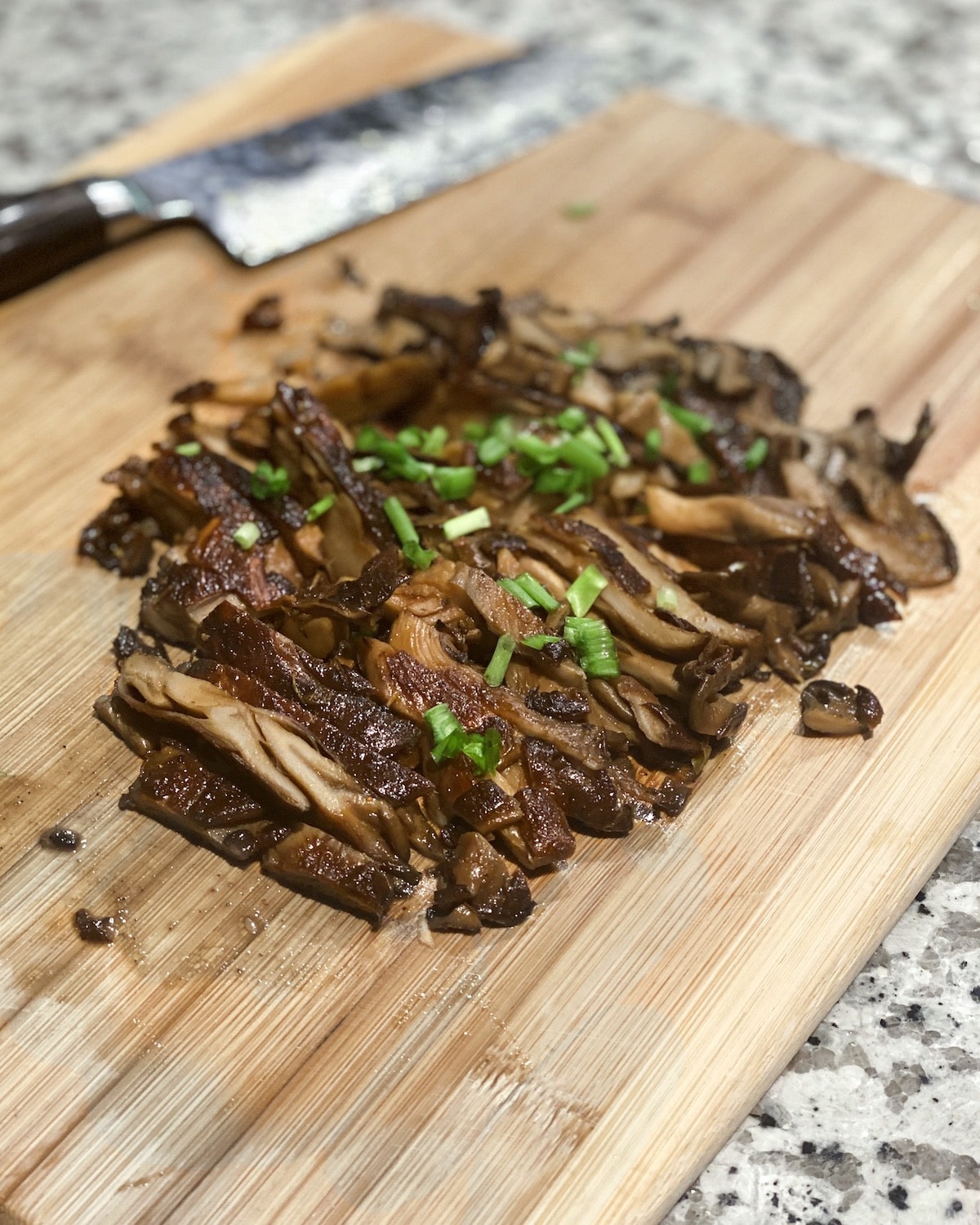 Teriyaki mushrooms garnished with spring onions on a brown bamboo cutting board with a knife in the background.